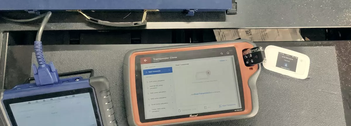 A key programmer mid-read on a transponder cloning screen, an OBD lead running off to the left and a smart fob resting beside it.