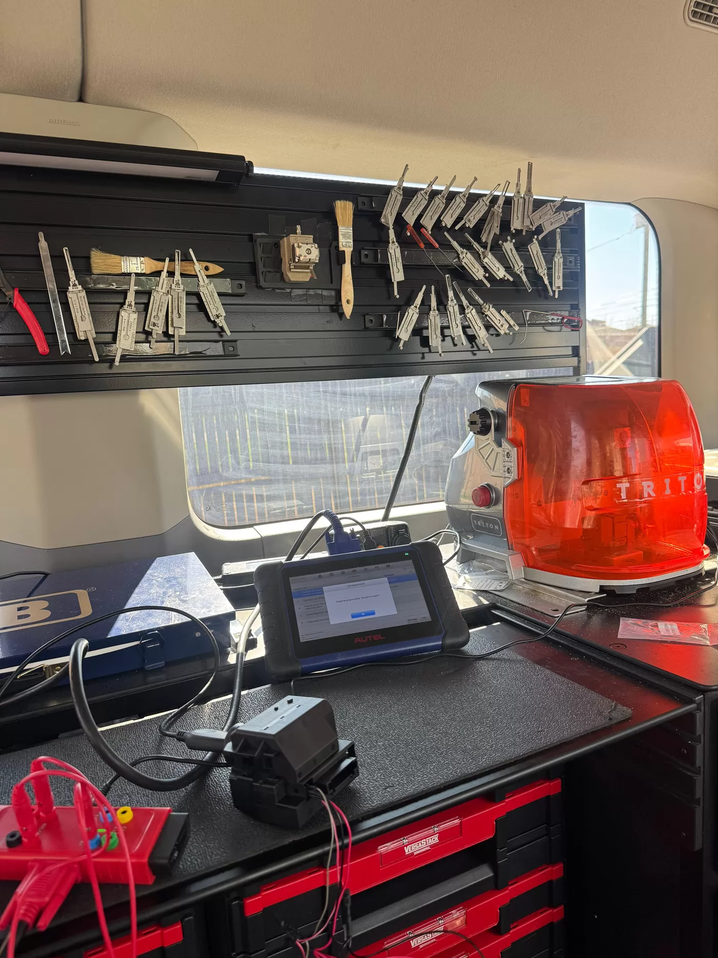 Workbench inside the service van: a rail of lock picks, a diagnostic tablet connected to an OBD lead, and a Triton key cutting machine.